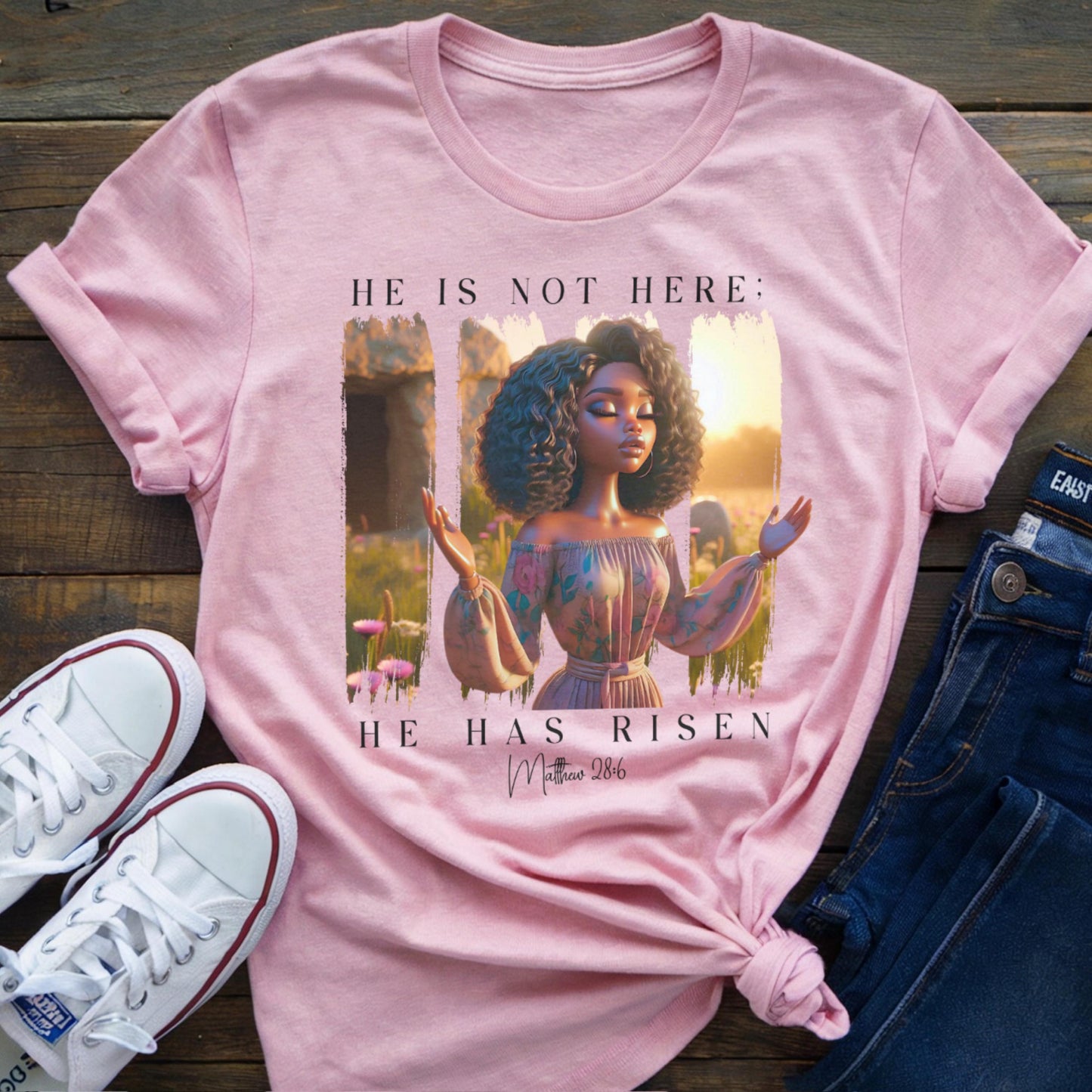 Pink t-shirt with a graphic of a woman and 'He Is Not Here' graphic on a wooden surface with shoes and jeans.