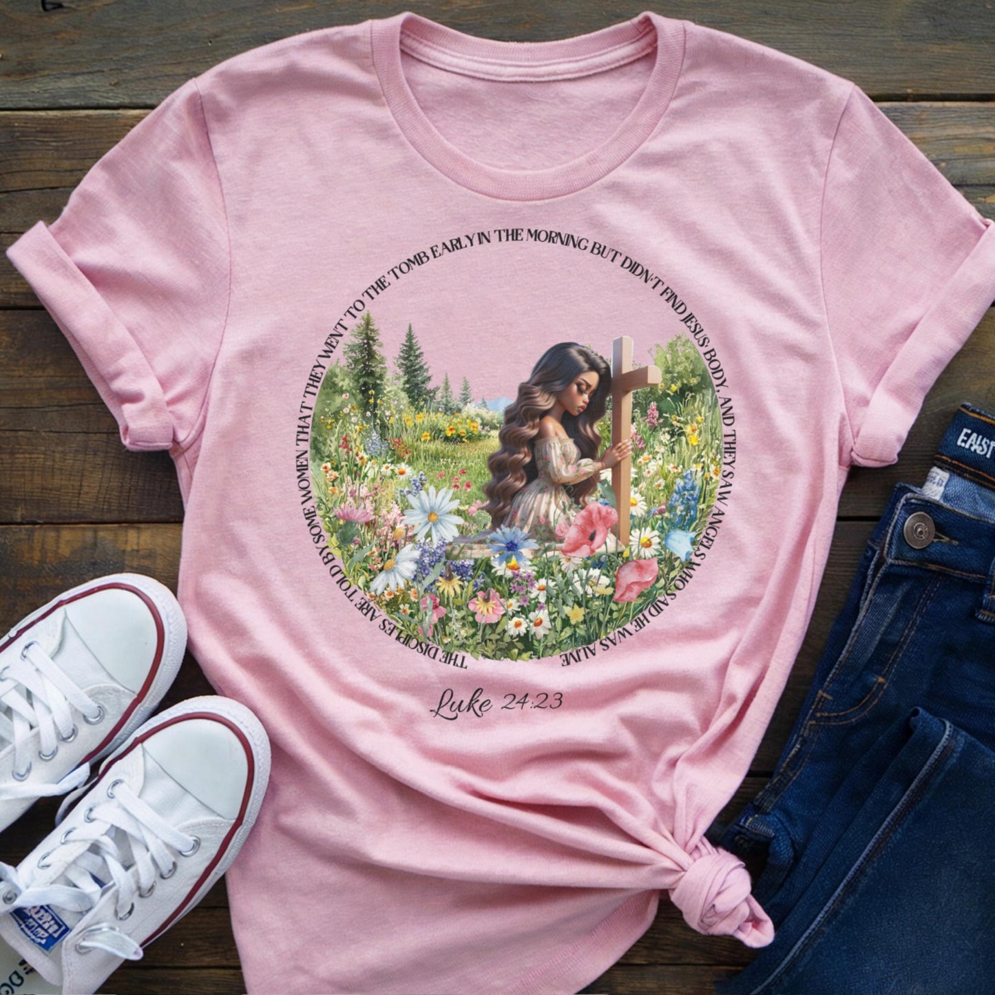 Pink t-shirt with a woman at a cross floral design, placed on a wooden surface with shoes and jeans.