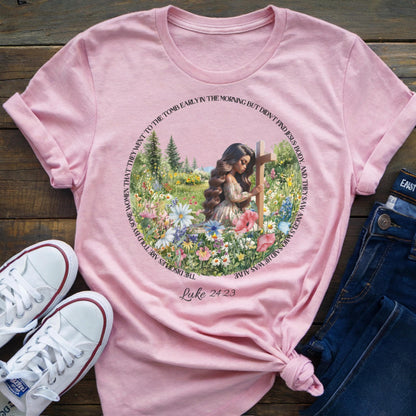 Pink t-shirt with a woman at a cross floral design, placed on a wooden surface with shoes and jeans.