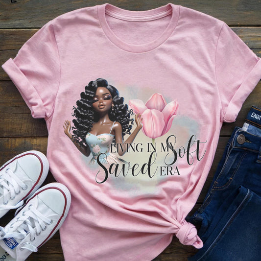Pink t-shirt with a graphic of a woman holding a pink flower and text, placed on a wooden surface with white sneakers and blue jeans.