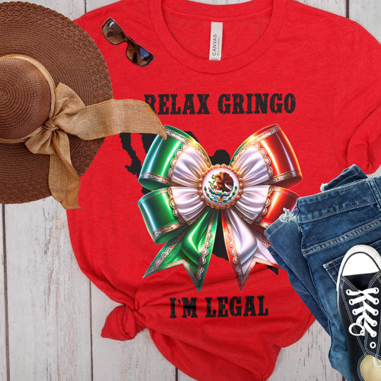 Red t-shirt with Mexico bow graphic and text 'Relax Gringo I'm Legal, accompanied by a straw hat, sunglasses, jeans, and sneakers on a wooden surface.