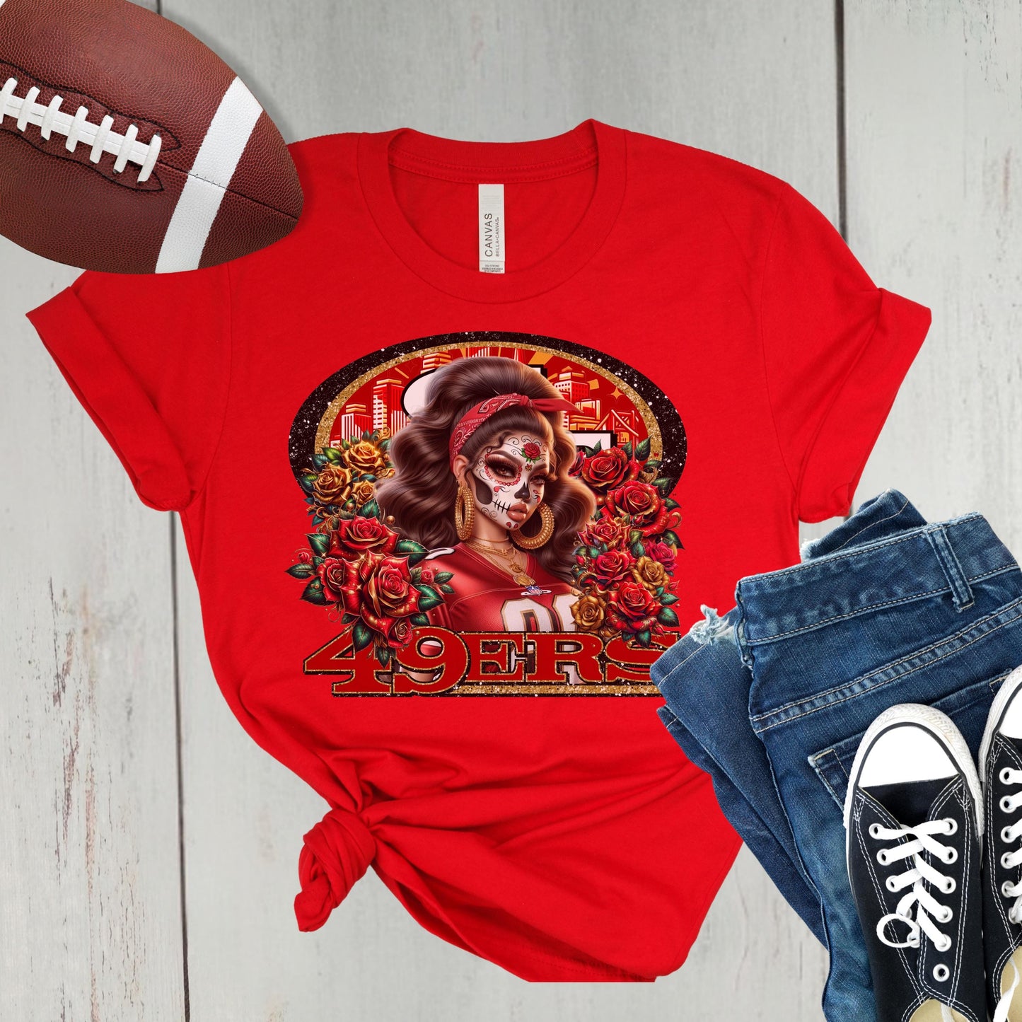 Red t-shirt with a dia de los muertos niners design, football, jeans, and sneakers on a wooden surface