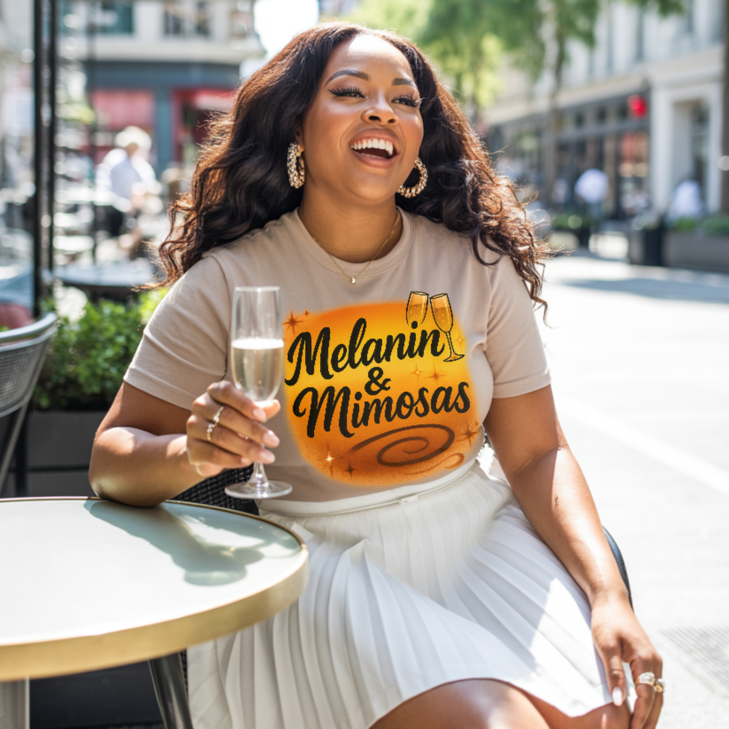 Woman holding a glass of champagne with 'Melanin & Mimosas' shirt in an outdoor setting