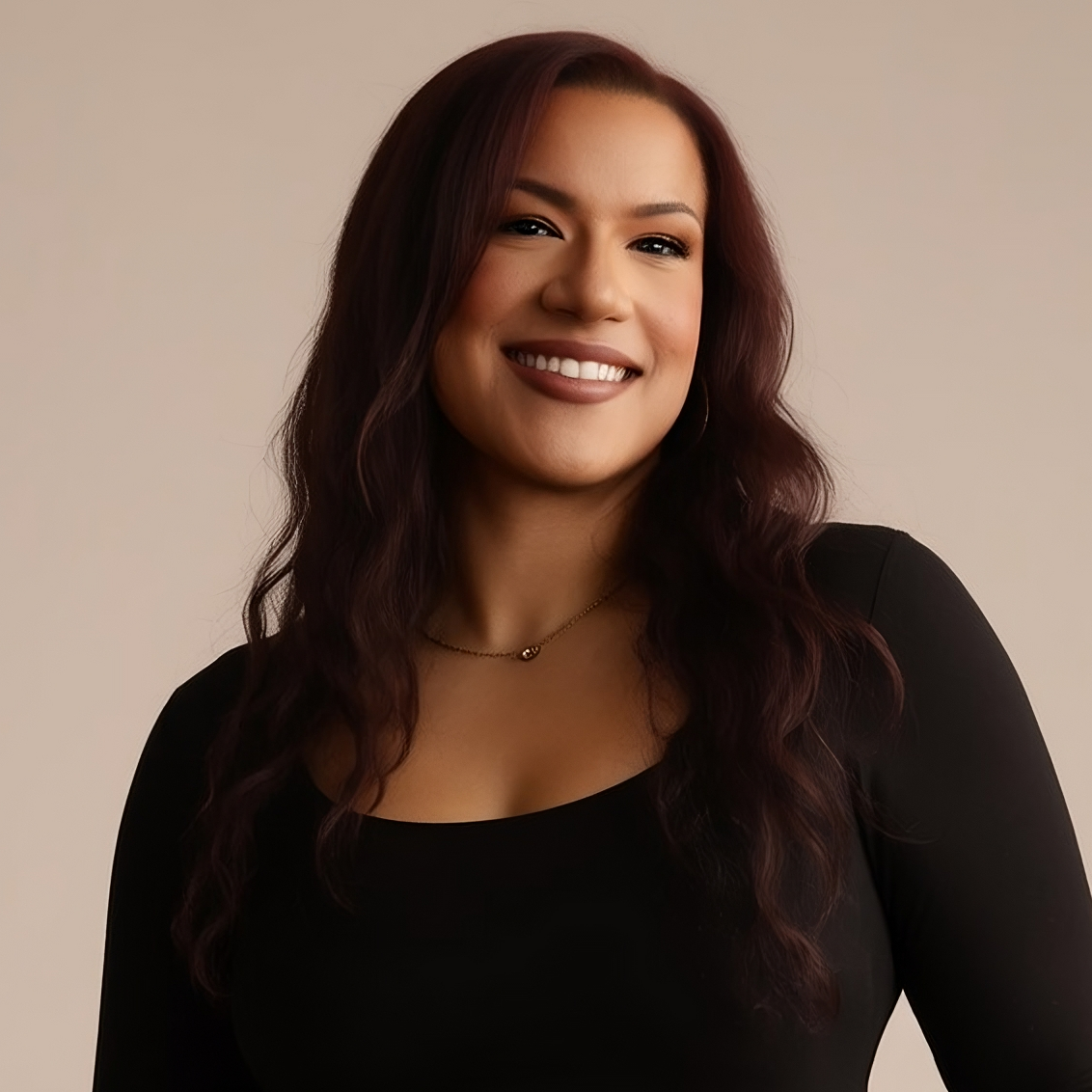 Woman wearing a black top and burgundy pants holding a black handbag against a neutral background