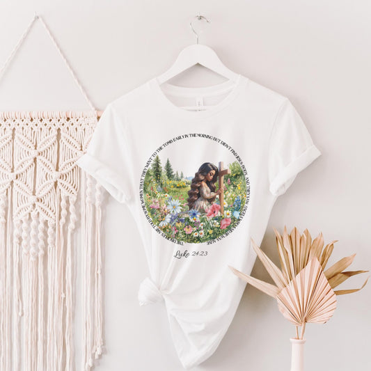 White t-shirt with a printed woman at a cross floral design on a hanger against a white wall.