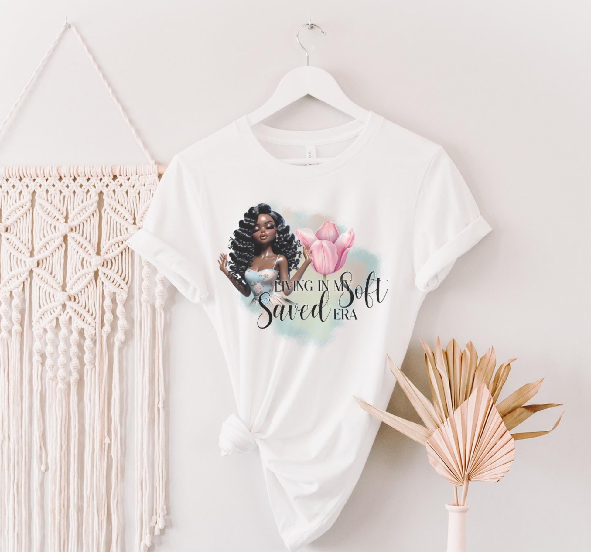 White t-shirt with a graphic design of a woman with braids and text, hanging on a white wall with decorative elements.