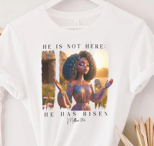 White t-shirt with a 'He Is Not Here' graphic and text on a hanger against a white wall.