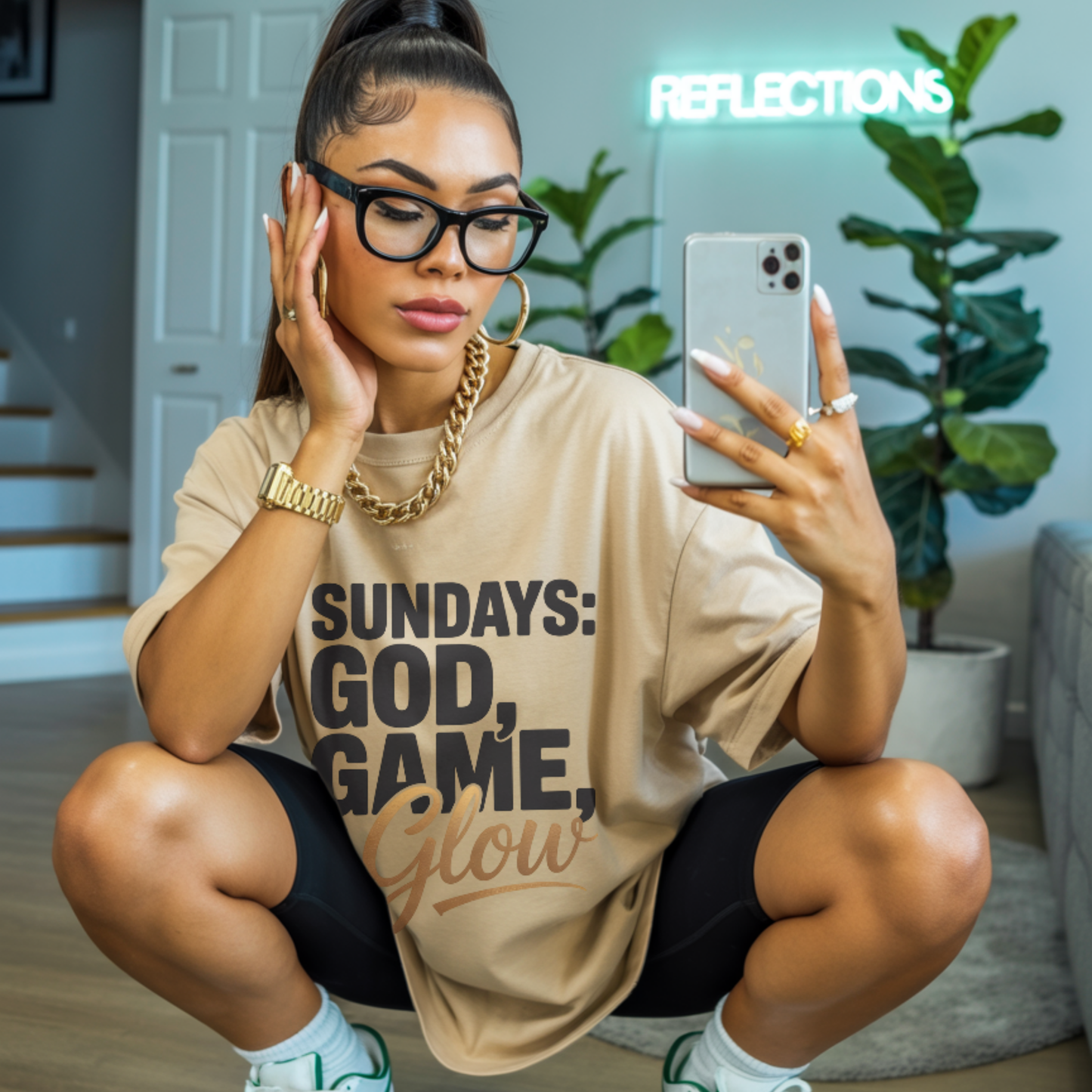 Woman taking a mirror selfie wearing a beige t-shirt with sundays god game glow design, sitting on the floor in a home setting.